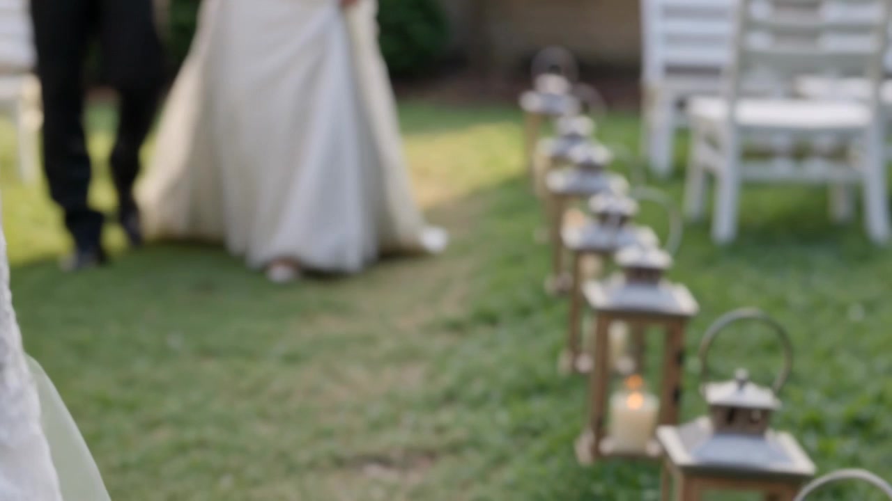 Sposa che cammina con bouquet in abito bianco lungo la navata erbosa mentre la cerimonia inizia, passando accanto a lanterne.