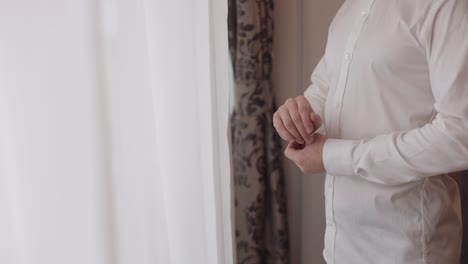 handsome groom man dresses in wedding morning and fixes his buttons on a white shirt sleeve, indoors