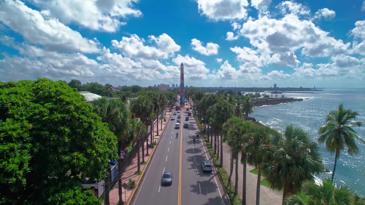 Ripresa aerea con drone dall'alto che si allontana e sale sopra il traffico di George Washington Avenue verso l'Obelisco Hembra, la costa e il Mar dei Caraibi a Santo Domingo