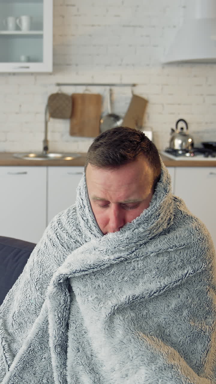 Man suffering from flu. Young sick man sitting on a sofa warming with blanket in the kitchen. Miserable man having a headache. Covid-19. Isolation at home. Vertical video