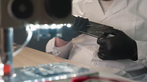 a close-up view of a technician in a white lab coat and black gloves examining a smartphone screen under a microscope