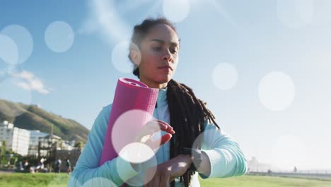 Mehrere-Weiße-Flecken-Schweben-Gegen-Eine-Fitte-Afroamerikanische-Frau,-Die-Auf-Dem-Sportplatz-Eine-Smartwatch-Benutzt