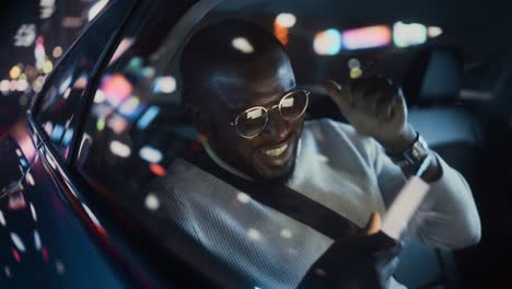 stylish black man in glasses is commuting home in a backseat of a taxi at night. handsome male playing video game on smartphone while in a car in urban city street with working neon signs.