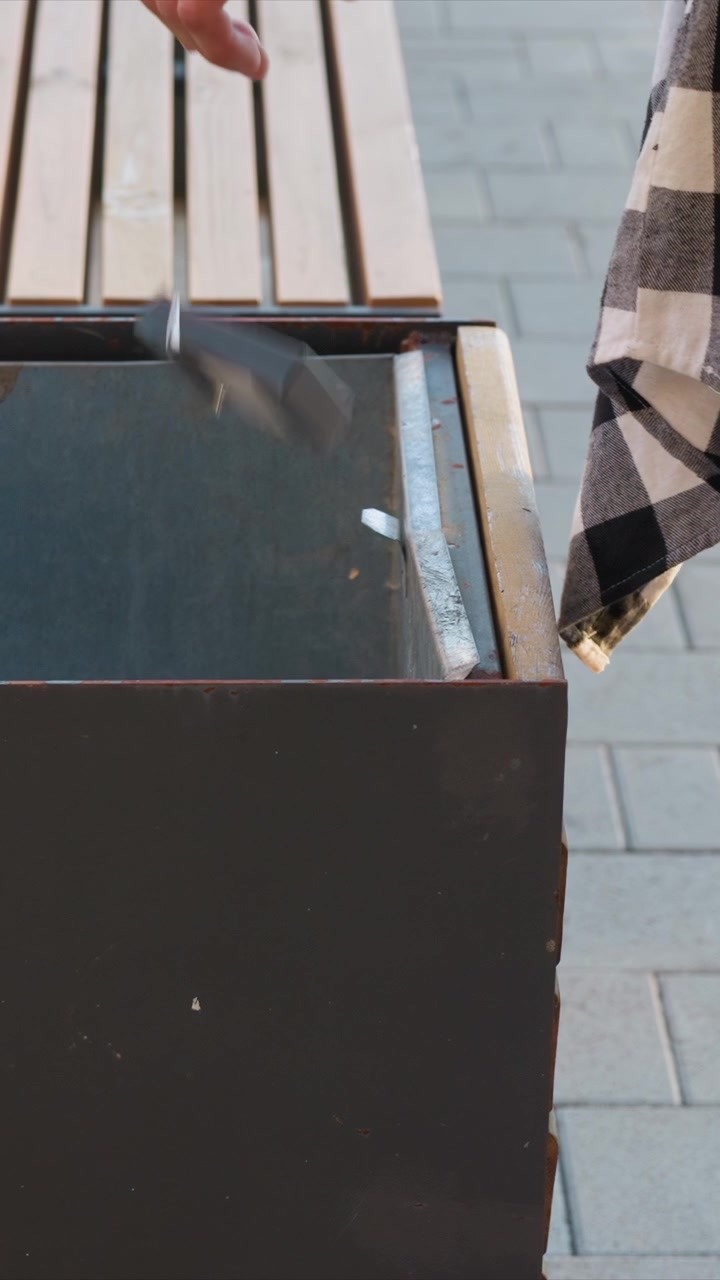 Close up of metal trash can with occasional small pieces of litter inside positioned next to wooden bench on gray brick pavement. Person wearing checkered shirt and jeans briefly appears