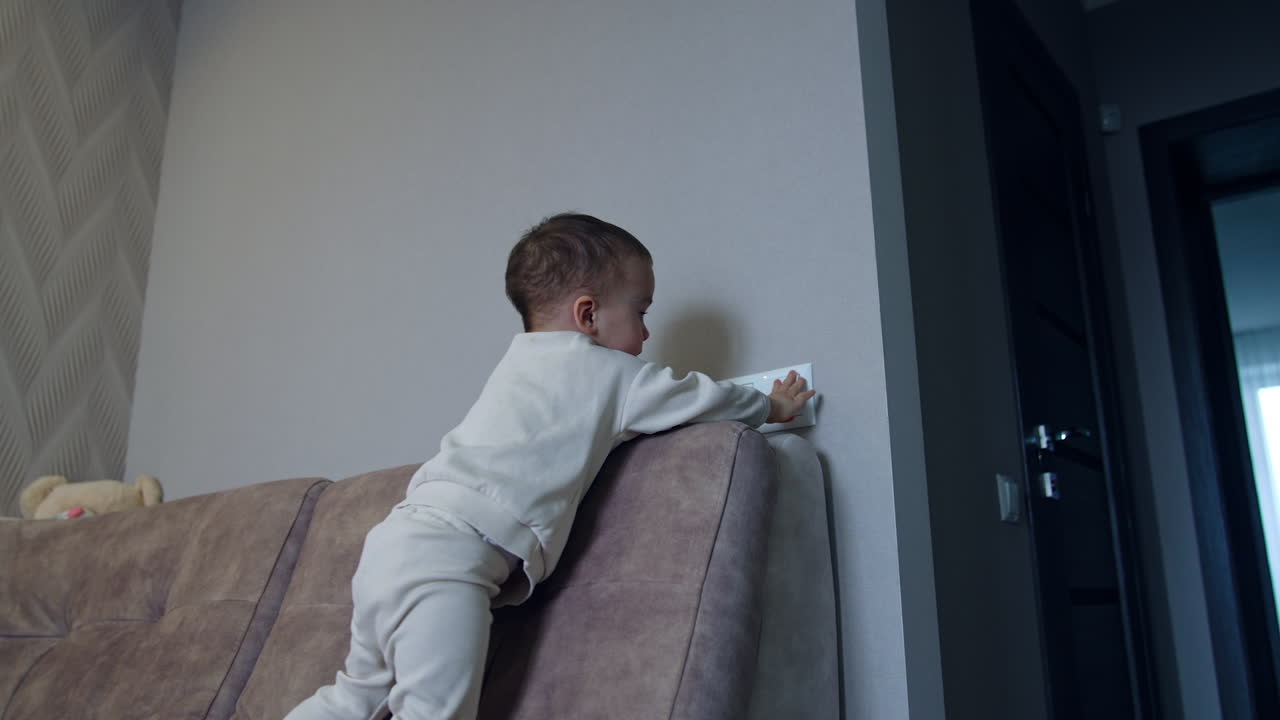Active kid walks by the sofa and reaches the light switch. Baby switches on illumination in the room. Low angle view.