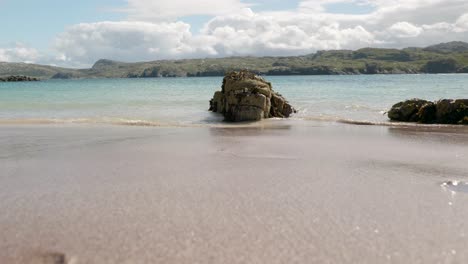 Die-Ebbe-Und-Flut-Einer-Meereswelle-Stürzt-An-Einem-Sandstrand-Stetig-Auf-Kristallklares-Meerwasser-über-Felsen,-Die-Mit-Blasentang-Algen-Bedeckt-Sind