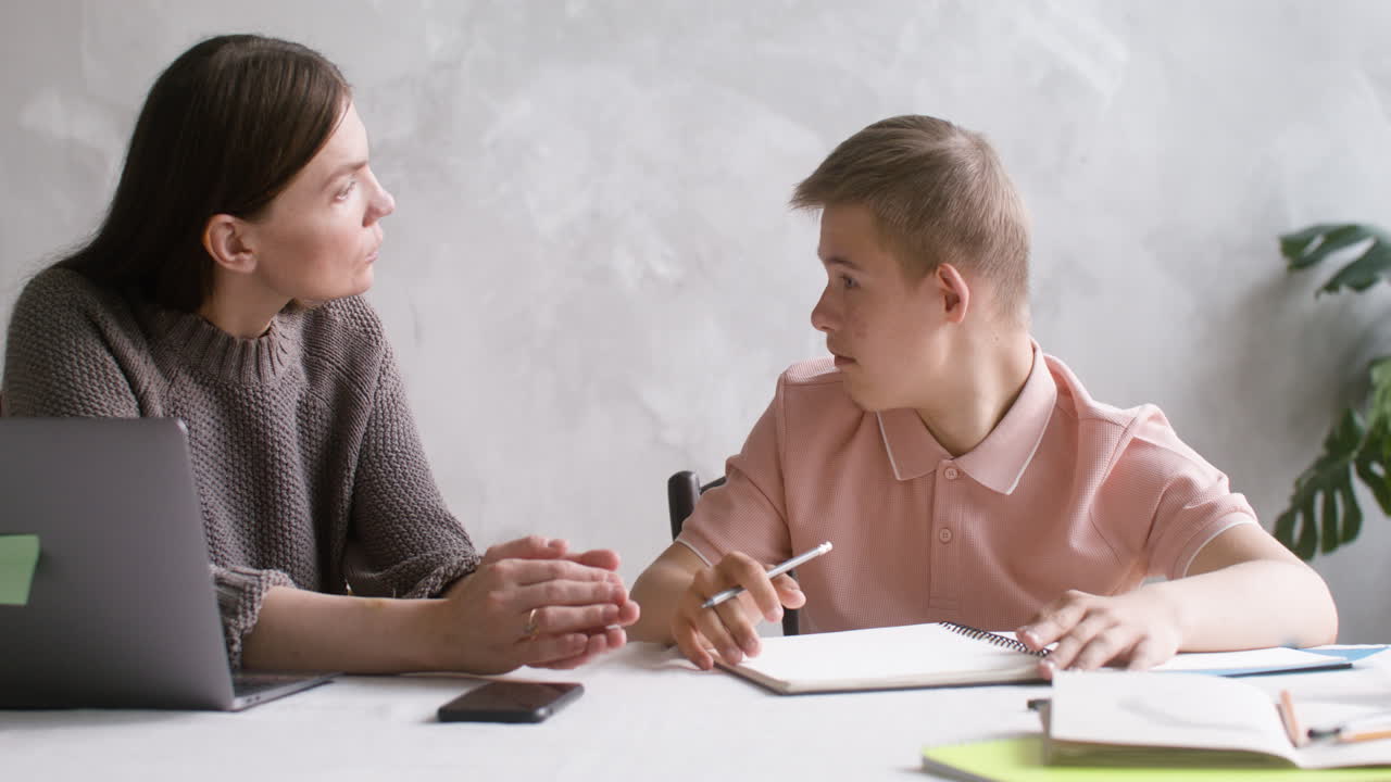 vista de cerca de un niño con síndrome de down haciendo tareas sentado en la mesa en la sala de estar en casa. su madre lo ayuda