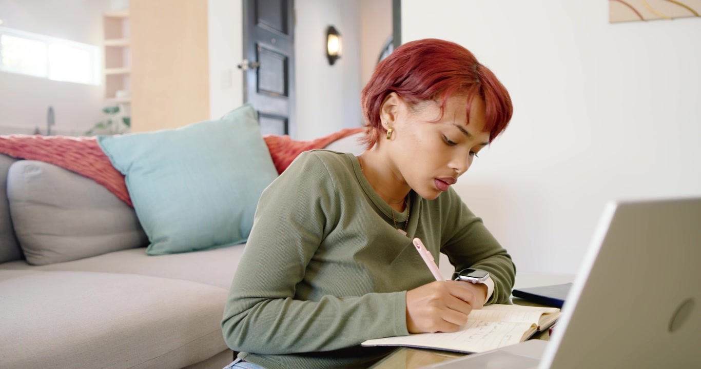 African woman sitting at low table glancing at laptop, taking notes, adjusting earbud while working