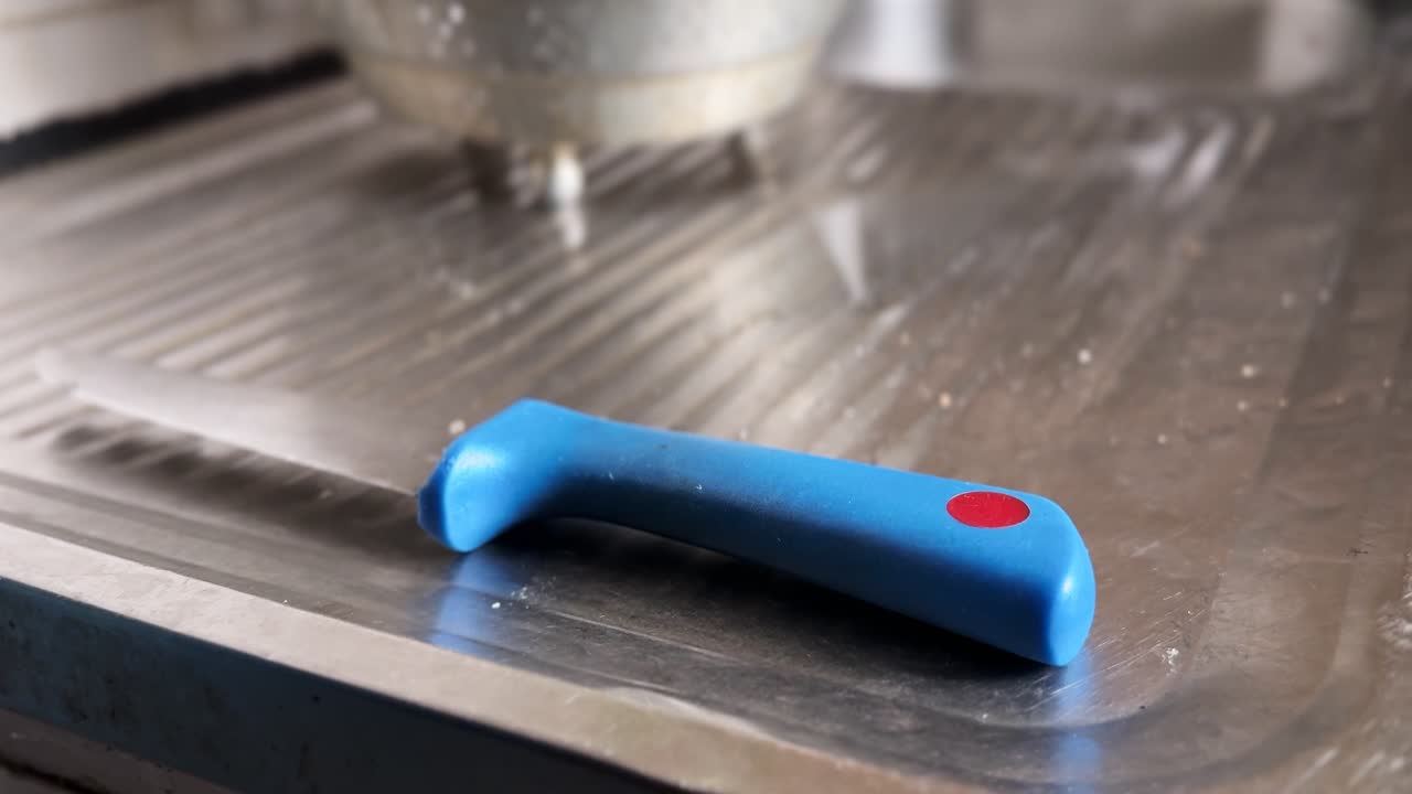 Primo piano del manico blu in plastica di un coltello da cucina con un pennarello rosso appoggiato su uno scolapiatti del lavello in acciaio inossidabile. Ambiente cucina interna che evidenzia i dettagli degli utensili e delle attrezzature per la preparazione del cibo.