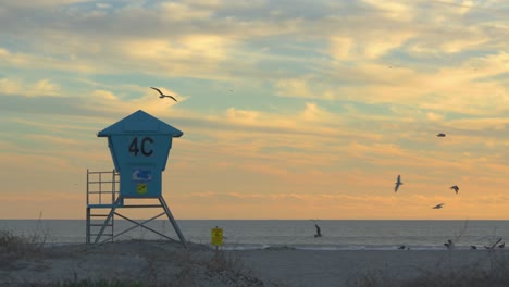 una torre de socorristas sentada en la playa durante una impresionante puesta de sol, mientras los pájaros vuelan alrededor