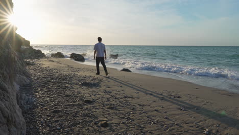 man-walks-on-beach-during-sunny-morning,-he-pauses-to-observe-beautiful-sea