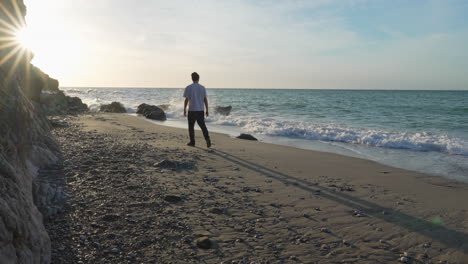 man walks on beach during sunny morning, he pauses to observe beautiful sea