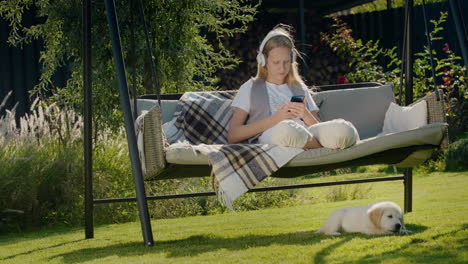 a teenage girl is resting on a garden swing, using a smartphone. her puppy sits next to her