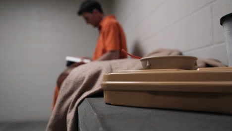 a prisoner in orange prison uniform, jumpsuit sits on his cot, bed reading