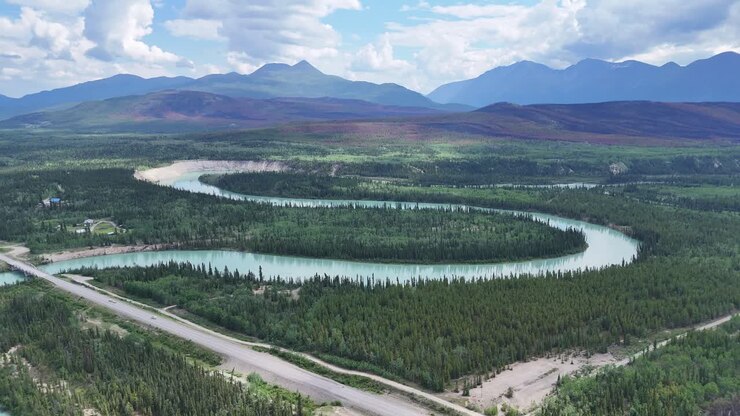 Un maestoso fiume turchese serpeggiante si fa strada attraverso una vasta foresta sempreverde con montagne blu distanti sotto un cielo nuvoloso dinamico in Alaska