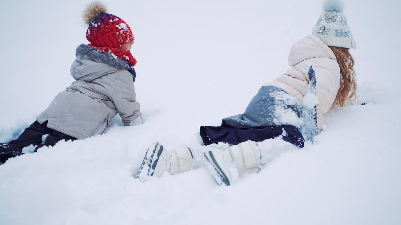 Back view of two kids crawling on snow on the white background in winter. Happy children are lying in soft snow outdoors. Close- up. Slow motion.