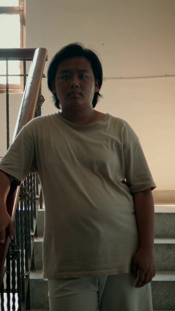 Video verticale: Adolescente maschio che nota la telecamera e si gira verso le scale tenendo il corrimano, indossando una maglietta chiara.