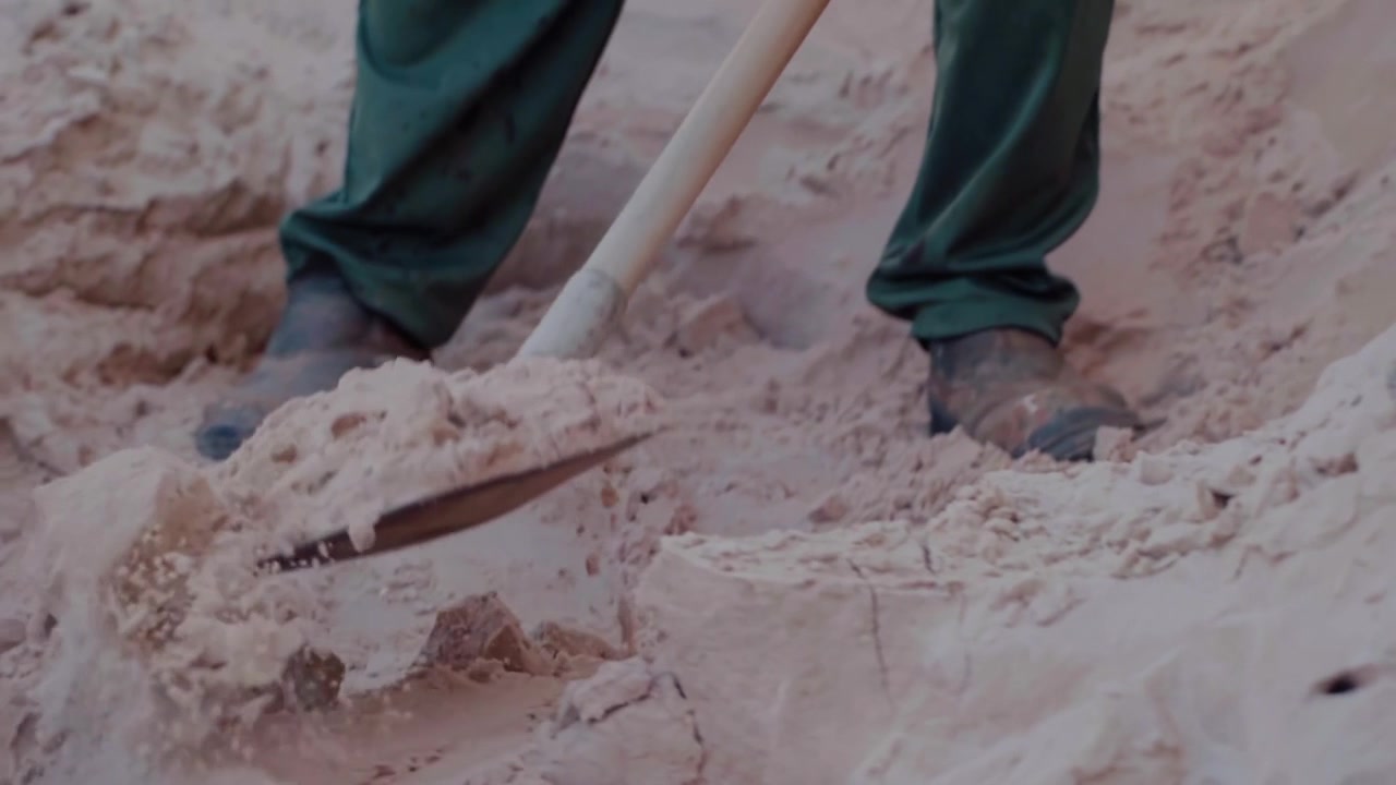 Close up of a worker's feet in work boots using a shovel to move sand. Manual labor on a construction site for sustainable building
