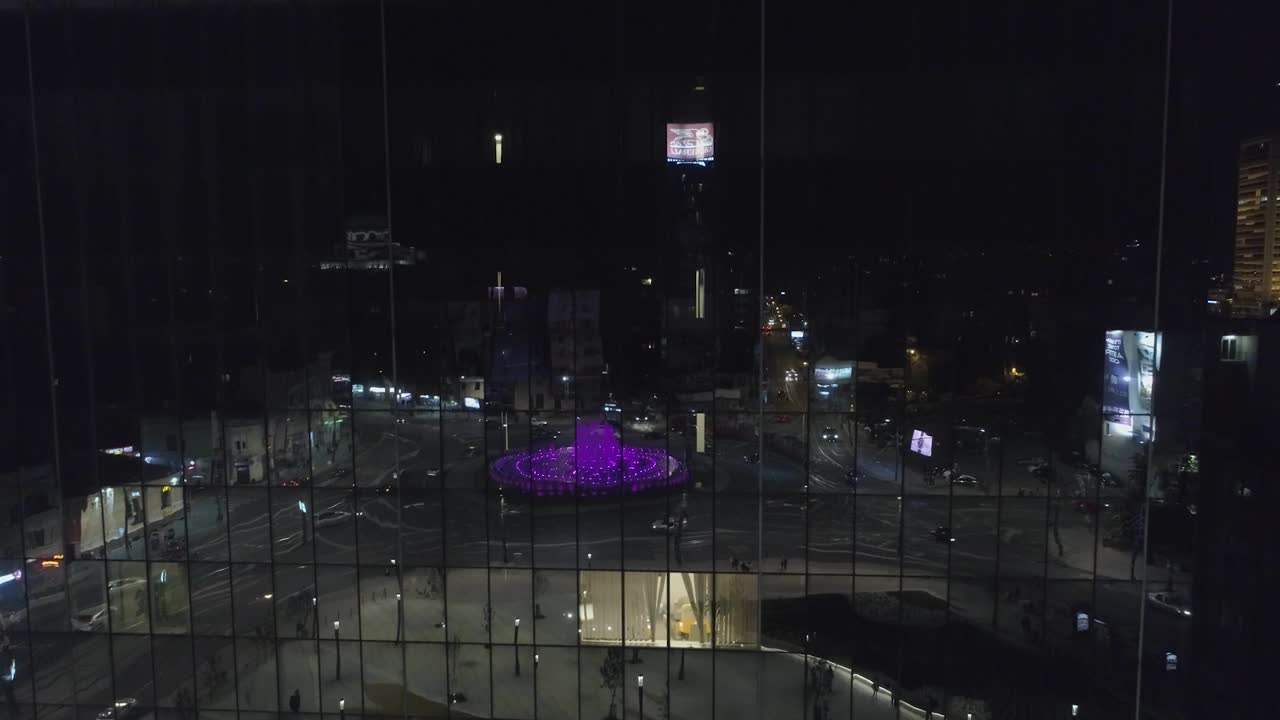 una toma aérea nocturna de alta definición que muestra el reflejo de una fuente colorida y una plaza de la ciudad en un edificio corporativo de vidrio