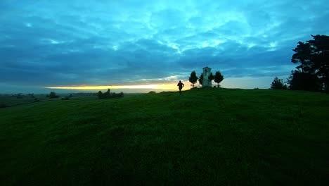 a teenager running up the hill and stopping at the top of the hill near a catholic shrine ,looking at a dramatic sunrise while the drone follows him and flies over his head