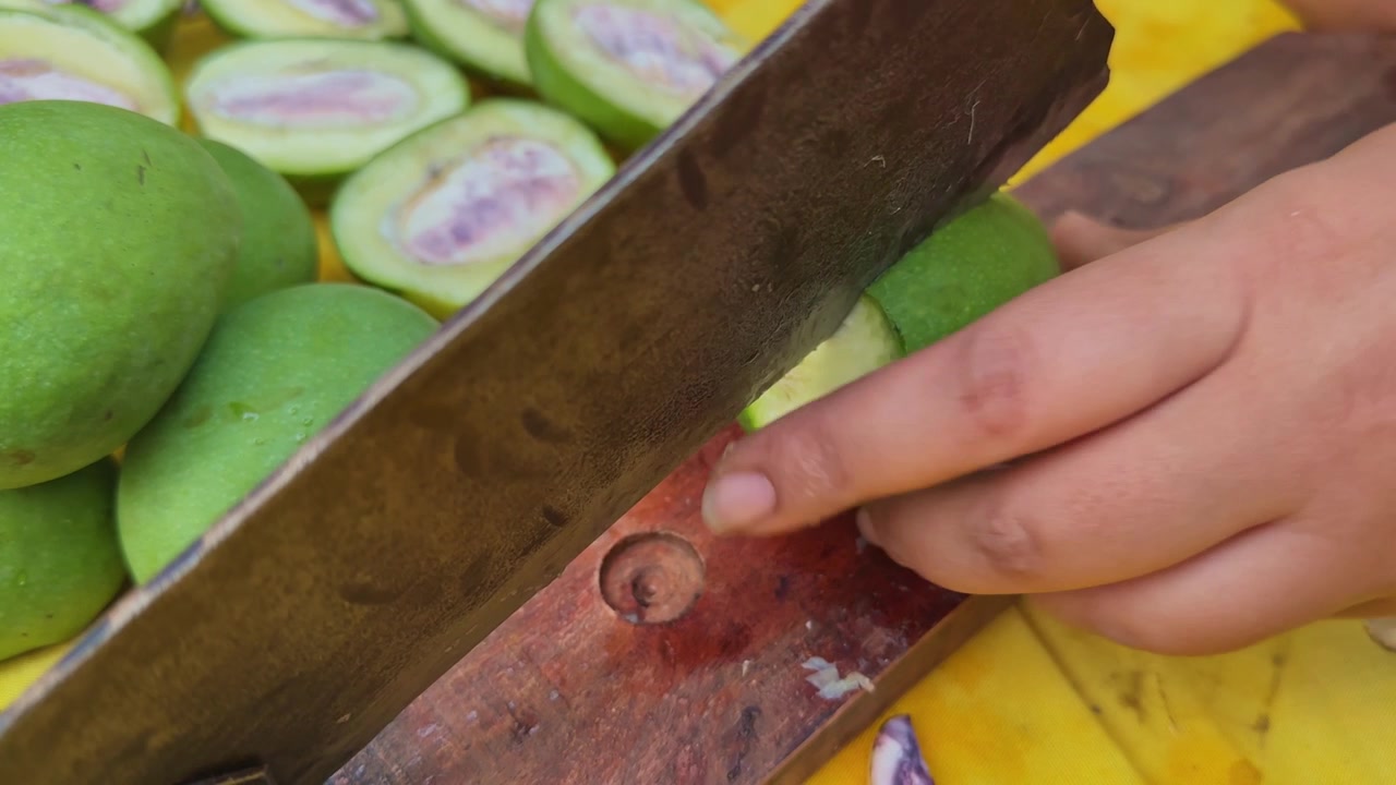 Vista ravvicinata dettagliata di mani che affettano manghi verdi crudi a metà con un tradizionale taglierino manuale indiano durante la preparazione casalinga di sottaceti su una superficie gialla.