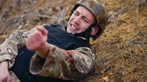 Nahaufnahme:-Ein-Soldat-In-Tarnuniform-Und-Schutzweste-Liegt-Mit-Einer-Verwundung-Am-Arm-Und-Winkt-Einem-Sanitäter-Zu,-Um-Bei-Kampfhandlungen-In-Der-Steppe-Erste-Hilfe-Zu-Leisten