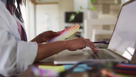 Mixed-race-woman-using-laptop-writing-in-notebook-drinking-coffee-working-from-home