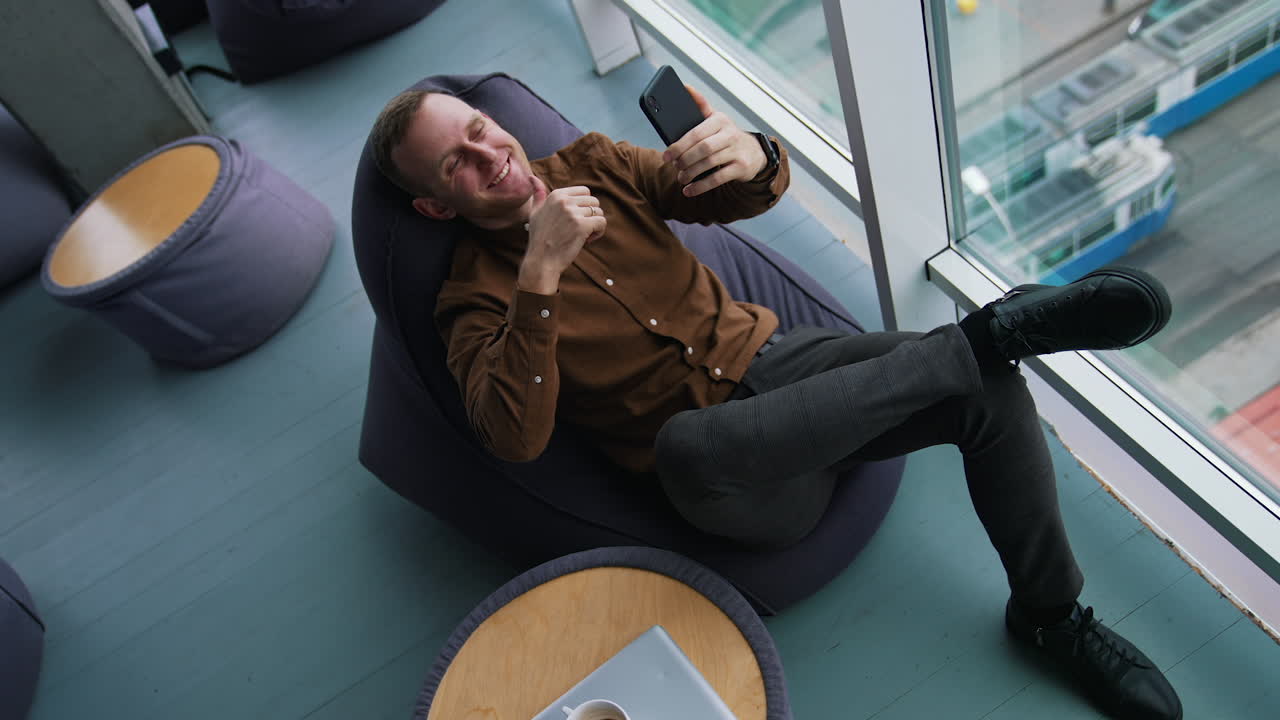 Businessman is having a video call. Young man in brown shirt and trousers sitting in a chair and talking online through the phone near the window. Top view.