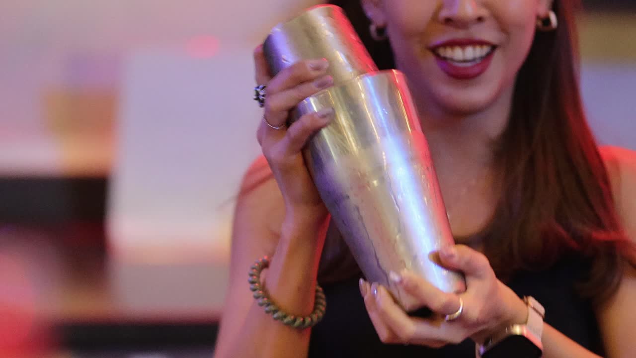Female bartender energetically shakes cocktail mixer, vibrant bar lighting, close-up, cheerful atmosphere, handheld camera
