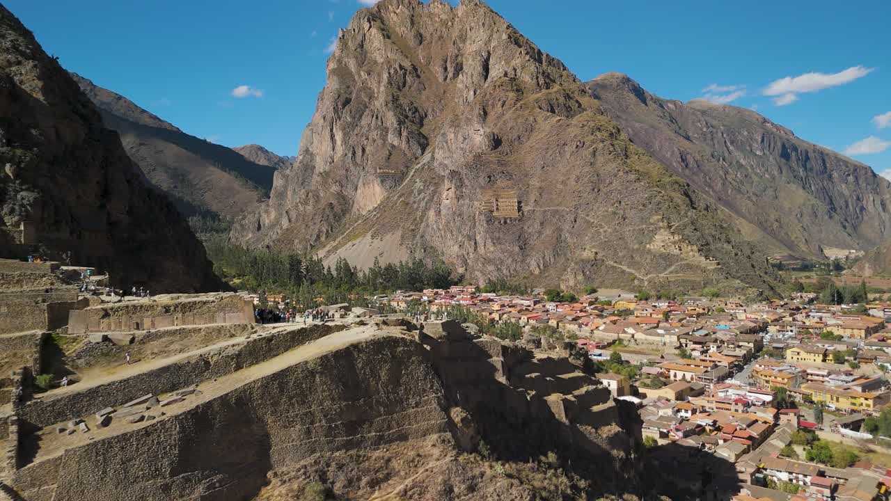 Wide drone shot of Ollantaytambo ruins and town below, surrounded by majestic Andean peaks under a clear blue sky