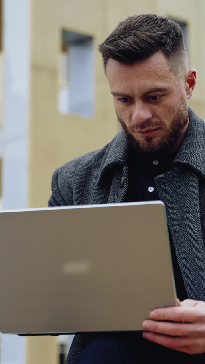 Portrait of a handsome man with a laptop. Young bearded businessman in a coat works on a wireless gadget on the urban background outdoors. Vertical video
