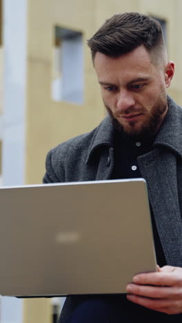 portrait of a handsome man with a laptop. young bearded businessman in a coat works on a wireless gadget on the urban background outdoors. vertical video
