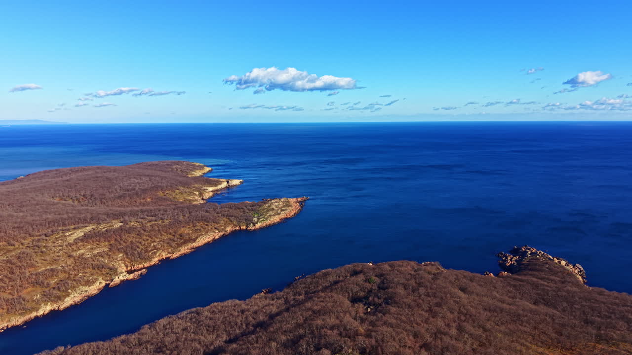 Kystlinjeutsikt over en steinete strand og klart vann om dagen under blå himmel med skyer nær et kupert område