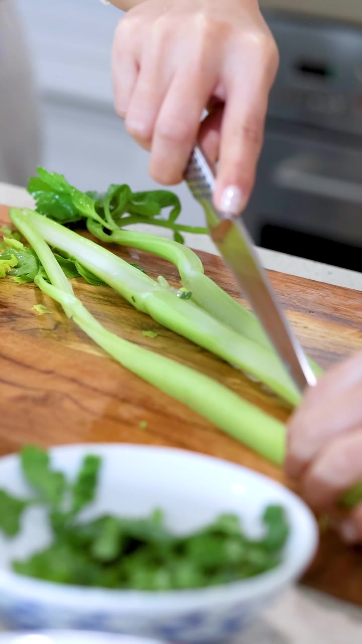 Mani che affettano il sedano su un tagliere di legno in una cucina luminosa. L'illuminazione naturale mette in risalto gli ingredienti freschi e l'ambiente pulito.
