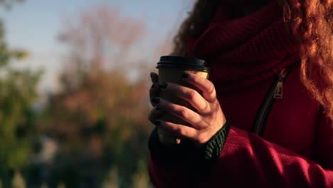 Alegre-Y-Elegante-Mujer-Joven-Con-Bufanda-De-Abrigo-Rojo-Parada-Al-Aire-Libre-En-La-Calle-Bebiendo-Café-A-La-Luz-Del-Sol-En-El-Parque-De-Otoño