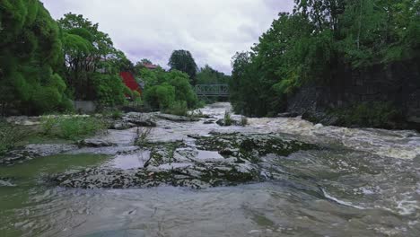 Flying-over-rapids-surrounded-by-trees-towards-a-bridge-in-Helsinki,-Finland