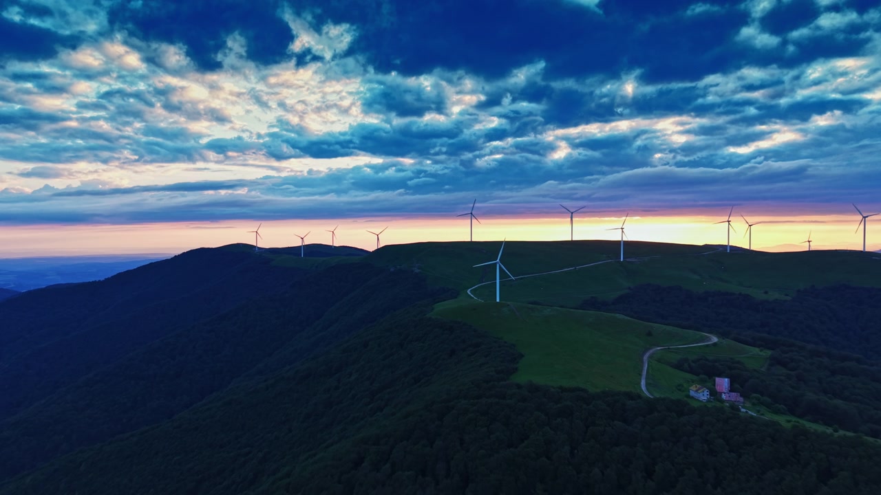 黃昏時分，日落餘暉灑在連綿山丘上，前景有風力發電機和農場，構成一幅風景如畫的景色。