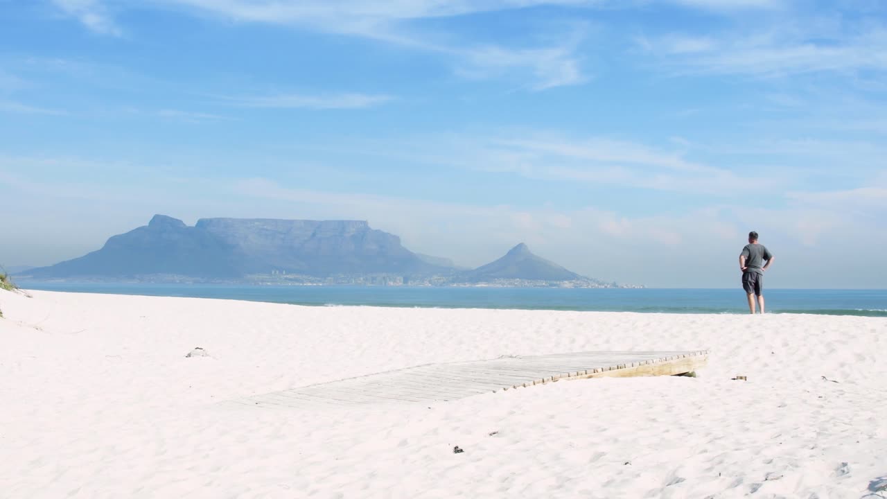 An international tourist jumps off a boardwalk onto Table Bay beach with Table Mountain in the background on a perfect Cape Town morning.