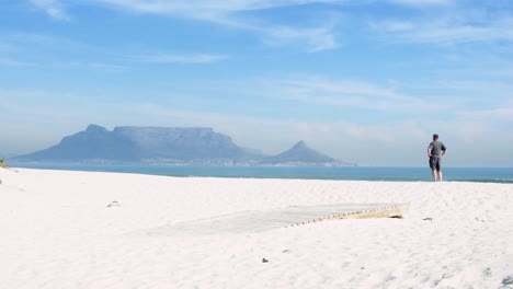 an international tourist jumps off a boardwalk onto table bay beach with table mountain in the background on a perfect cape town morning.