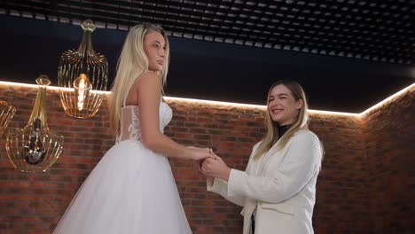 two women holding hands in an interior, one wearing a wedding dress