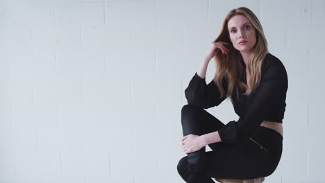 retrato de moda de una mujer joven seria sentada contra la pared del estudio blanco