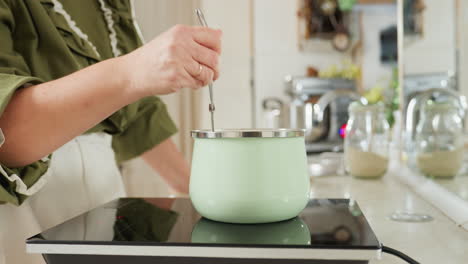 woman in green top and white apron stirs food inside pastel green pot on electric stove in bright modern kitchen, focusing on precise hand movement with metal utensil