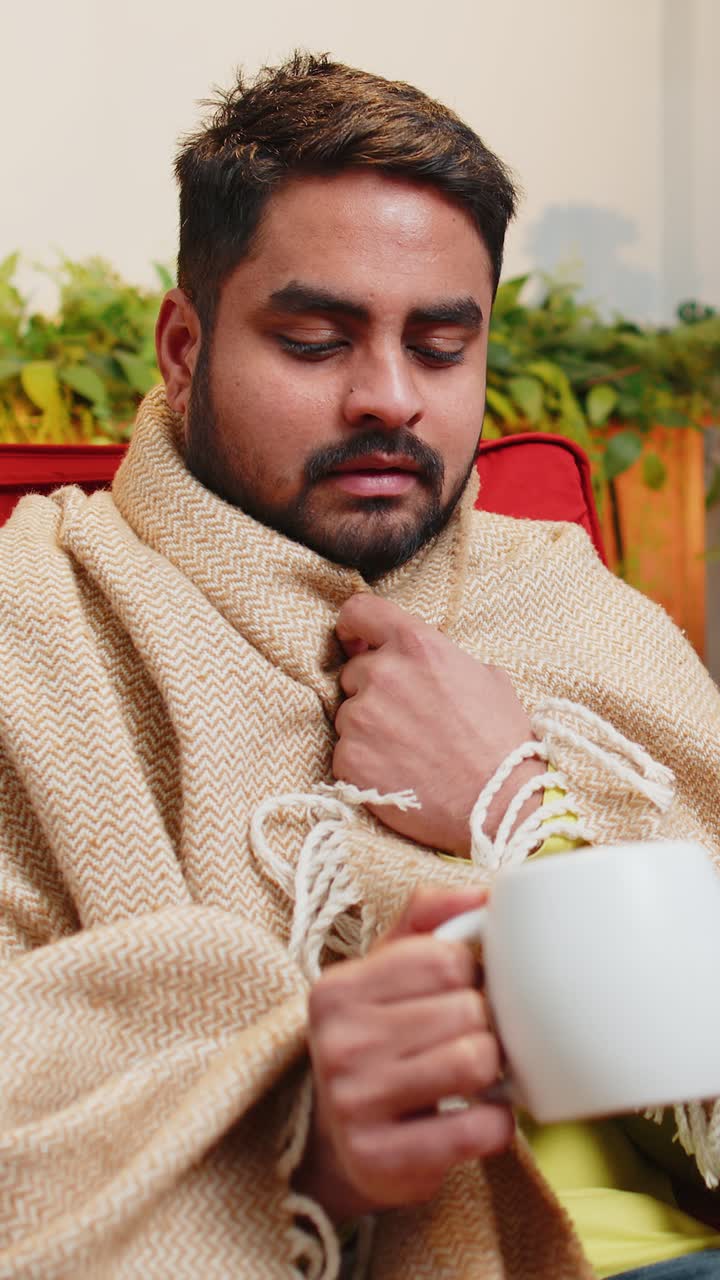 Sick indian man wrapped in plaid sit alone shivering trembling from cold on chair drinking hot tea