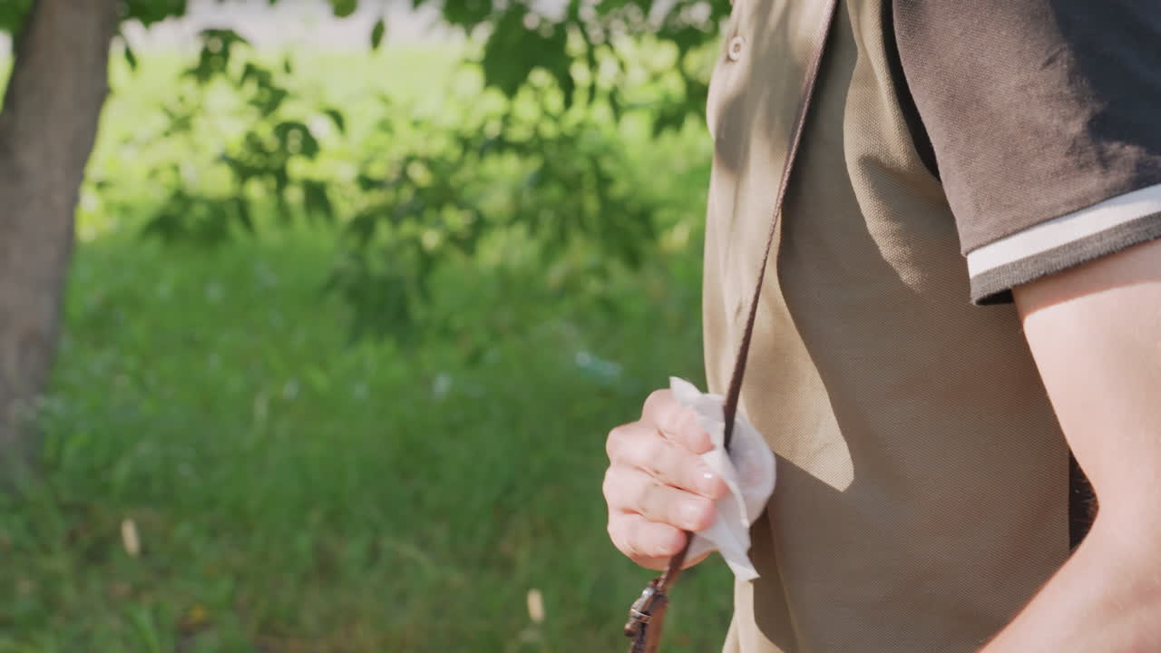 Young Man Dabbing Bag Strap With Tissue In Park Shade, Repetitive Blotting Action, Careful Palm Movements, Casual Polo Shirt And Sling, Warm Grassy Background, Focused Closeup Of Hands