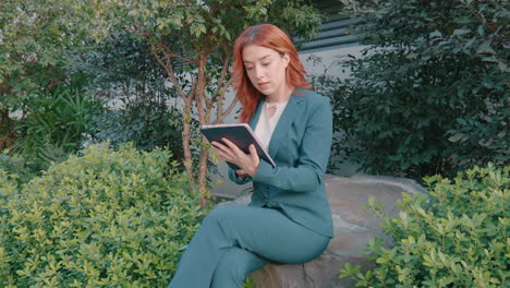 Shot-of-a-caucasian-woman-sitting-on-a-rock-in-a-park-and-using-a-tablet-to-scroll-through-her-office-work-on-a-sunny-day