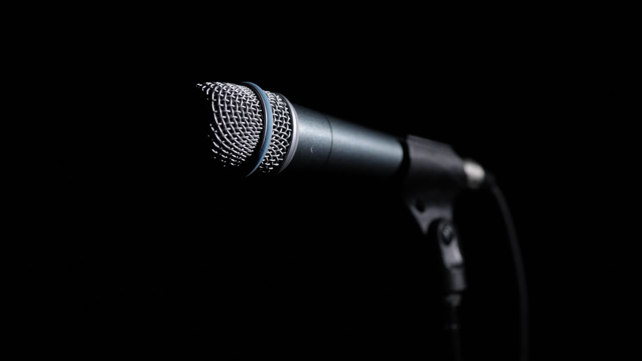 Dynamic microphone stands in profile against pitch black background with dramatic side lighting hitting silver mesh grille