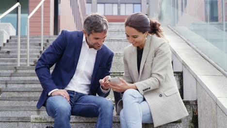 Businessman-And-Businesswoman-Sitting-By-Steps-Having-Meeting-Outdoors-Looking-At-Phone