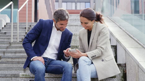 businessman and businesswoman sitting by steps having meeting outdoors looking at phone