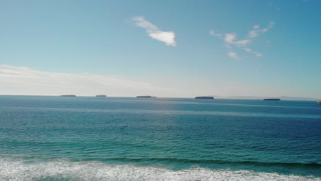 Many-cargo-ships-parked-off-the-coast-of-The-Port-of-Los-Angeles,-low-view-with-waves-in-foreground