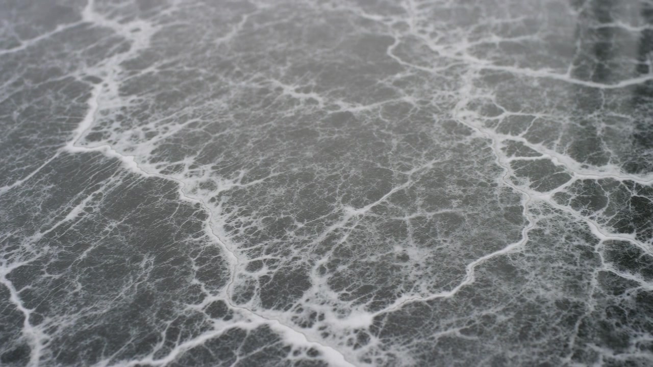 Bovenaanzicht van een donkere leigrijze zee bedekt met verweven witte schuimaders en schuimige filamenten, die delicate rimpelingen, organisch contrast en een kalme monochrome oppervlaktetextuur vormen.